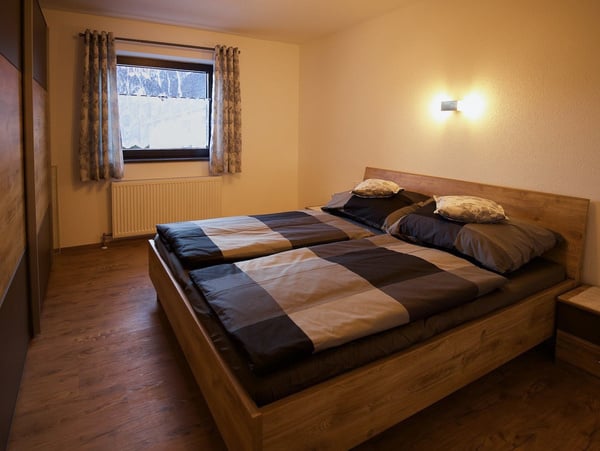 Modernes Schlafzimmer mit Holzbett und grauschwarzer Bettwäsche. Große Fenster mit gemusterten Vorhängen, Holzboden und Wandheizung. Dezente Wandbeleuchtung schafft eine gemütliche Atmosphäre. Perfekt für stilvollen Komfort und erholsamen Schlaf.