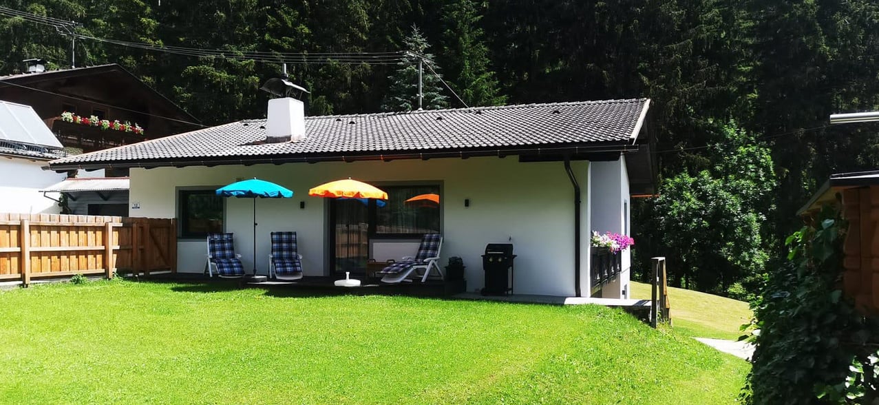 Einladendes Haus im Sommer, umgeben von grüner Wiese und Wald. Weiße Fassade mit Terrasse, ausgestattet mit bunten Sonnenschirmen und Liegestühlen. Ideal für entspannte Tage im Freien und Grillabende. Perfekte Lage für Naturgenießer.