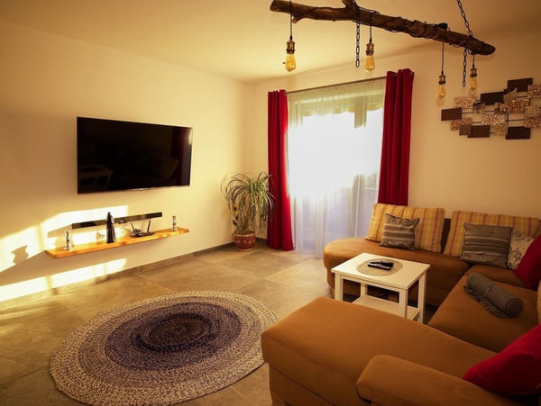 Modernes Wohnzimmer mit L-förmigem braunem Sofa, roten und gestreiften Kissen. Flachbild-TV an der Wand, Holzregal, einzigartige Hängelampen, roter Vorhang und runder Teppich. Sonnenlicht durchflutet den Raum. Ideal zum Entspannen.