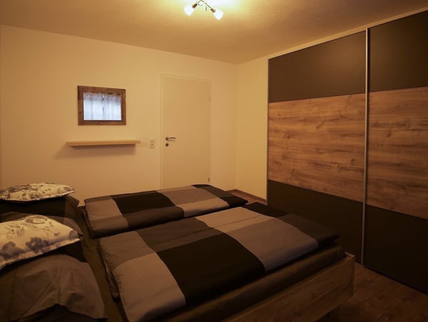 Modernes Schlafzimmer mit einem großen Holzbett und grau-schwarzer Bettwäsche. Großer Einbauschrank mit Holz- und Schwarzakzenten, schlichte Wanddekoration und dezente Beleuchtung. Perfekt für komfortablen und stilvollen Schlaf.