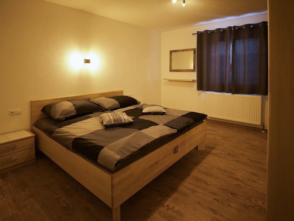 Gemütliches Schlafzimmer mit großem Holzbett, moderner grauer und schwarzer Bettwäsche, ambienter Beleuchtung und dunklen Vorhängen. Holzboden, Nachttisch und Wandheizkörper vervollständigen den minimalistischen Raum.