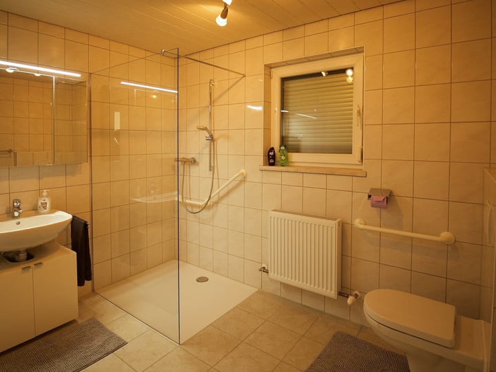 Modernes Badezimmer mit begehbarer Dusche, großen Fliesen und viel natürlichem Licht. Ausgestattet mit einem Waschbecken, Spiegel und einem Fenster für Belüftung. Handtuchhalter und Heizkörper sorgen für Komfort. Elegantes und funktionales Design.