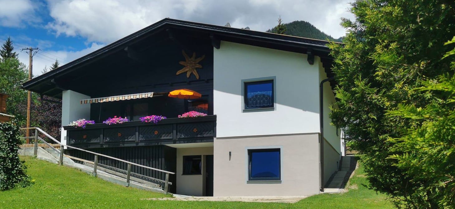 Charmantes Alpenhaus mit schwarz-weißer Fassade, großem Balkon mit bunten Blumen und orangefarbenem Sonnenschirm. Umgeben von üppigem Grün und Bergen, bietet es eine malerische und ruhige Atmosphäre. Ideal für eine erholsame Auszeit.