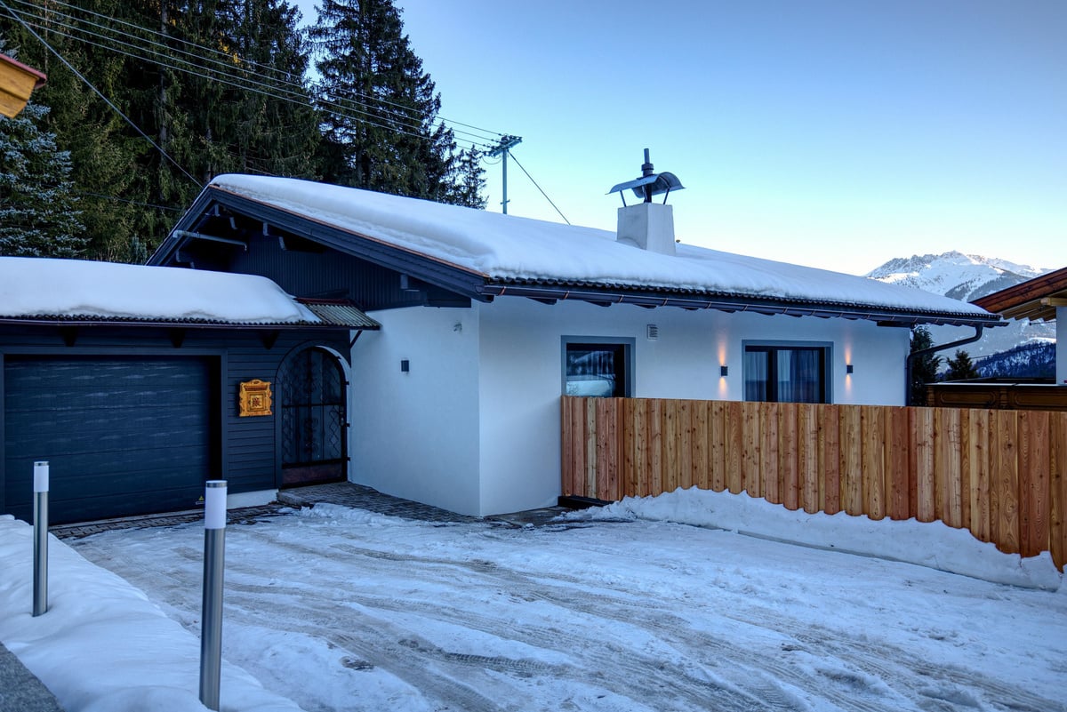 Charmantes Haus im Winter, umgeben von Schnee und Bergen. Weiße Fassade mit modernen Fenstern und Holzzaun. Überdachter Eingang und Garage bieten Schutz vor den Elementen. Ideale Lage für eine gemütliche Winterauszeit in den Alpen.